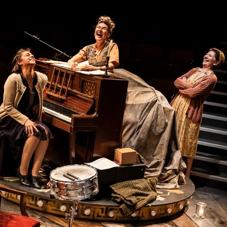 Group of women in 1940s clothes laughing around a piano