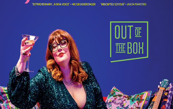 A woman with ginger hair, wearing glasses and a green sequin dress sat on a sofa with a martinin glass held up in the air