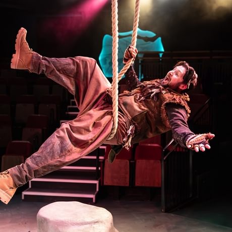 A man in a brown costume lying on a rope swing with pink and yellow lights in the background.