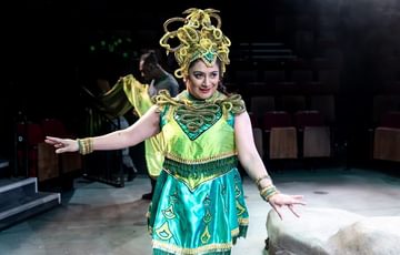 A woman in a crown with a man holding a long piece of green fabric in the background. With white lights around them.