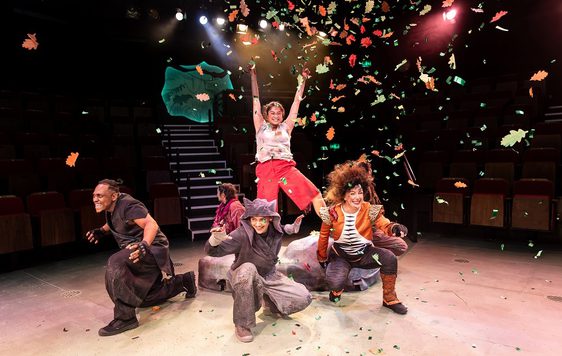 A woman standing on a rock with her hands up. Five people squatting around the rock with orange and green confetti flying around them and yellow, white, and red lights in the background.