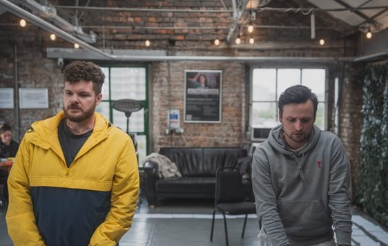 Two men looking sad. Man 1 has short curly hair and a beard he is wearing a yellow and man 2 has straight short hair and is wearingblue jacket and the other is wearing a grey jumper