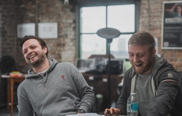 Two men sat at a table laughing, both wearing grey jumpers.